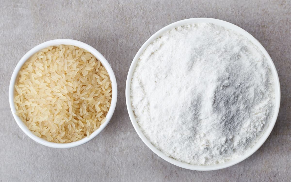 Raw rice grains and flour in white bowls on kitchen table